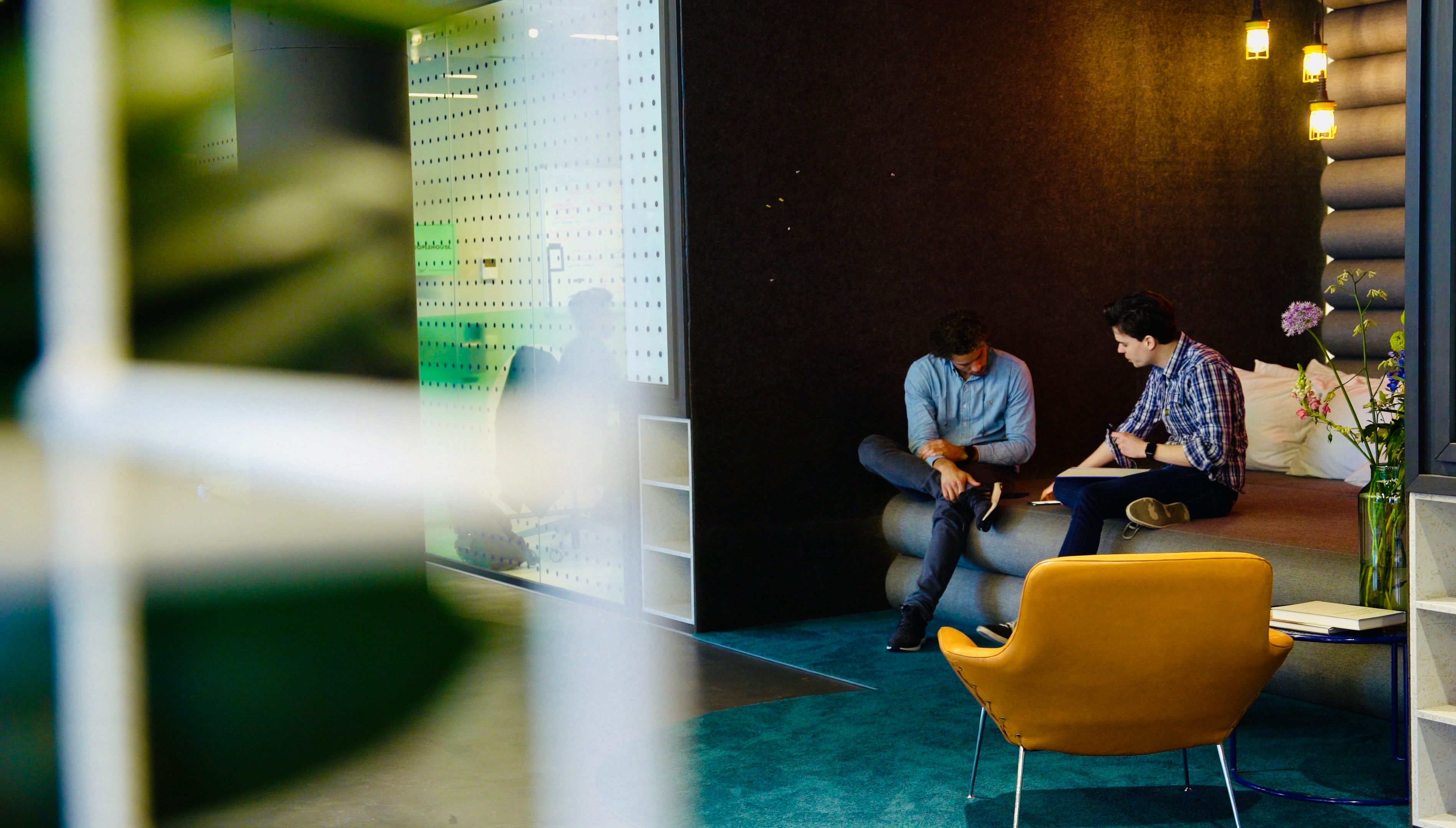 A conversation in a workspace with teal carpet, warm lighting and a yellow chair.